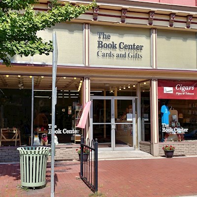 Tan building with glass windows and doors. Toys, books, and clothing can be see for sale through the windows. The Book Center cards and gifts is written over the door. There are red bricks in front of the building.