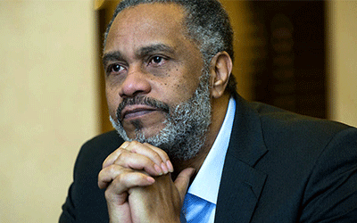 Anthony Ray Hinton resting his chin on his clasped hands. He is wearing a black suit. 