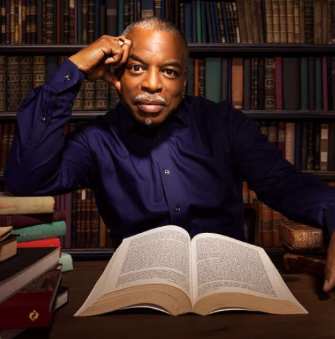 LeVar Burton sitting in front of bookshelves, an open book in front of him, with his head resting on his hand as he looks at camera