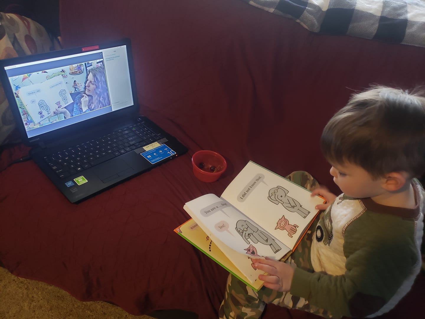A Story Time friend watching virtual Story Time on a laptop while sitting on the couch with an open book