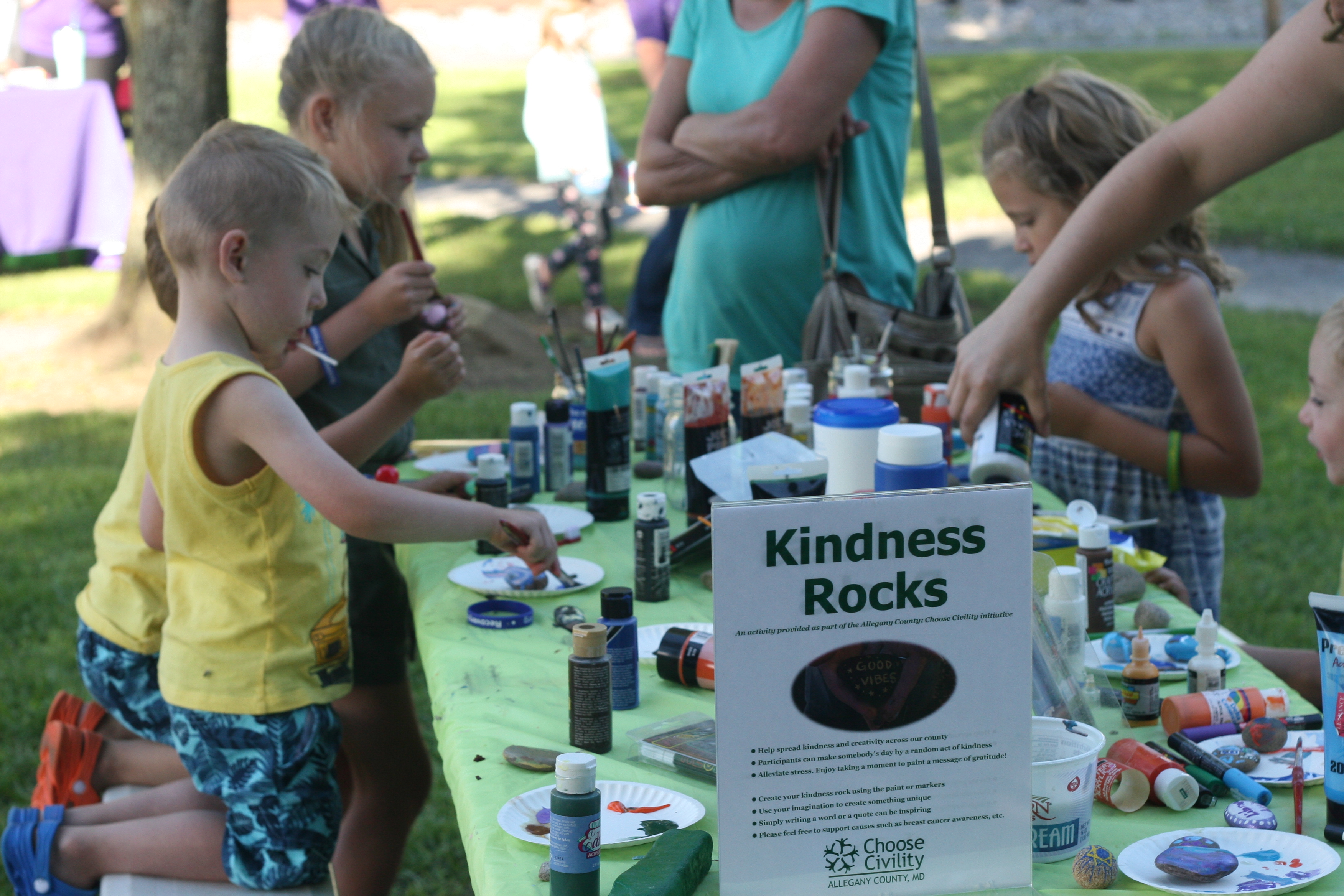 Table surrounded with kids and adults painting rocks. Sign labeled kindness rocks